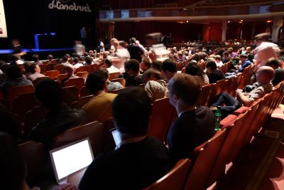 The photo shows the audience at the dConstruct conference, lots of seated people in the room, some of them with their laptops open; all are looking at the stage where the â€˜dConstructâ€™ logo is displayed